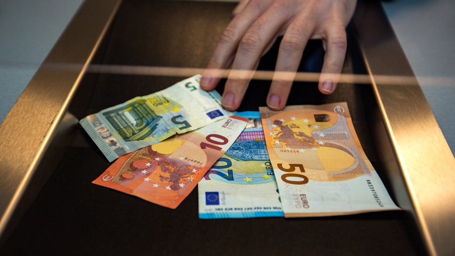 Eine Hand legt mehrere Geldscheine in einen Behälter, der an den Seiten silber glänzt. Der Behälter sieht aus wie die Durchreiche an einem Bankschalter. Es sind Scheine im Wert von 50, 20, 10 und 5 Euro. 