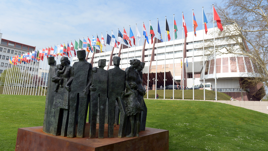 Auf einem Sockel auf einer Rasenfläche steht eine Skulpturengruppe mit sieben Personen. Im Hintergrund des Kunstwerkes steht ein modernes Gebäude mit einer auffälligen Rotunde an der Seite. Vor dem Gebäude befinden sich Flaggenmasten mit Nationalflaggen.
