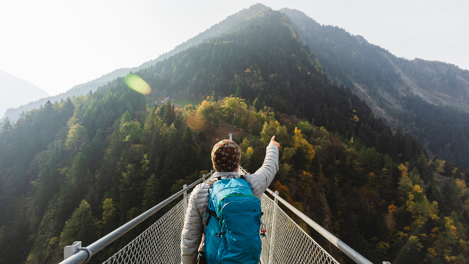 Ein Wanderer mit blauem Rucksack befindet sich in den Bergen auf einer Hängebrücke und zeigt mit dem rechten Arm in Richtung der vor ihm liegenden Gipfel. Die Hänge um ihn herum sind bewaldet. Die Sonne scheint. 