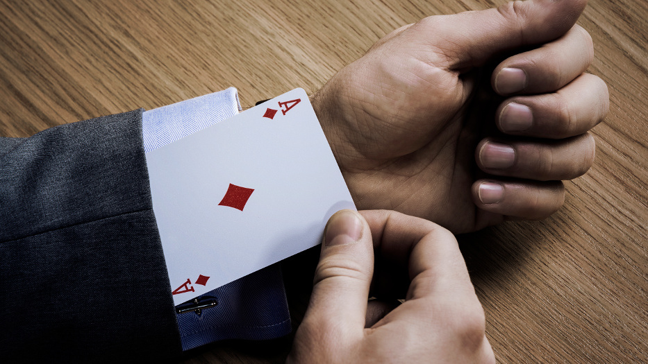 Eine Person, von der nur der Unterarm und die Hand auf einer Holz-Oberfläche zu sehen sind. Die Person trägt ein dunkles Jacket und ein weißes Hemd, aus dem Ärmel zieht sie eine Spielkarte mit einem roten Ass (Karo). 