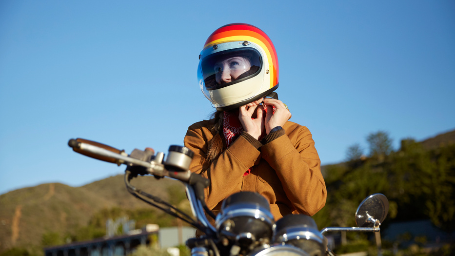 Eine Frau mit einer hellbraunen Jacke befindet sich an ihrem Motorrad und bindet sich ihren Helm fest. Der Helm ist unten sandfarben und hat über dem Visier ein auffälliges Regenbogen-Muster. Im Hintergrund sind Hügel und blauer Himmel zu erkennen.